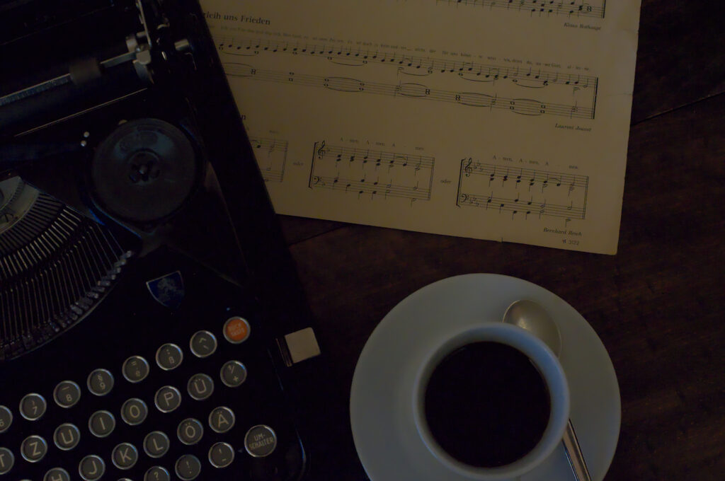 Auf einem Holztisch steht eine Schreibmaschine, ein Notenblatt und eine Tasse Kaffee
